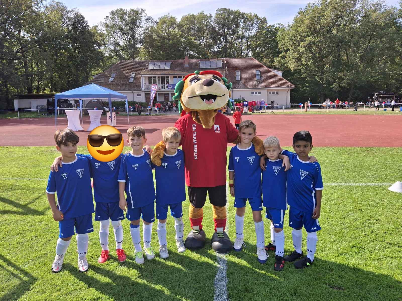 Sechs junge Fußballspieler von Blau-Weiss Spandau stehen gemeinsam mit einem Vereinsmaskottchen auf dem Fußballplatz. Die Kinder tragen blaue Trikots und posieren vor dem Vereinsgelände bei strahlendem Sonnenschein.